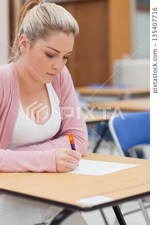 Female student sitting on blue chair at desk in classroom writing on paper with orange pen Female student sitting on blue chair at desk in classroom writing on paper with orange pen 135407716