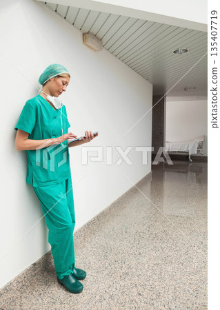 Female nurse in scrubs with mask leaning against wall using tablet in hospital corridor, copy space Female nurse in scrubs with mask leaning against wall using tablet in hospital corridor, copy space 135407719