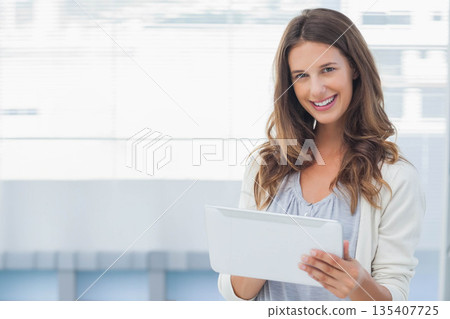Female professional in office attire standing at window in office holding tablet device, copy space 135407725