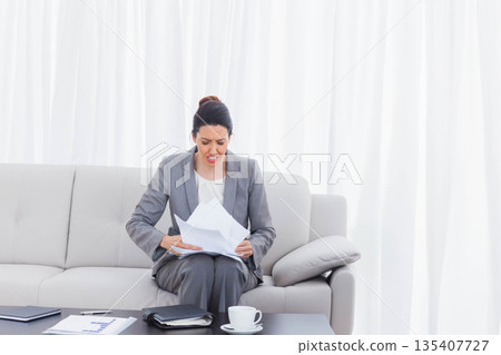 Businesswoman wearing suit sitting on sofa in lounge holding papers and grimacing near coffee cup 135407727