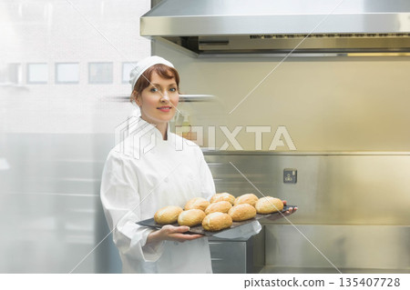 Female chef wearing chef coat holding tray of golden rolls under stainless hood in kitchen 135407728