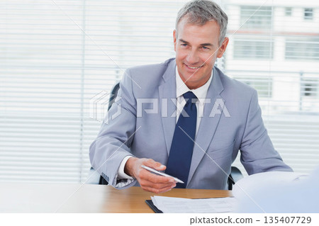 Senior man holding pen over stack of documents at wooden desk by window blinds in office 135407729