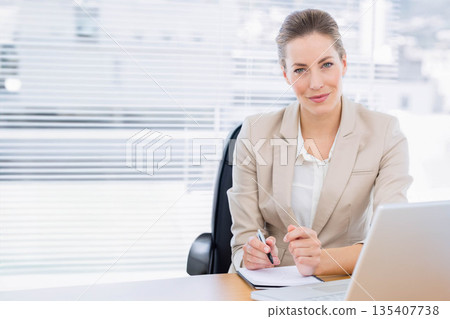 Businesswoman wearing blazer writing with pen in notebook at office desk with laptop, copy space Businesswoman wearing blazer writing with pen in notebook at office desk with laptop, copy space 135407738