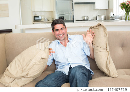 Male laughing while holding cushions on couch in living room with kitchen appliances and flowers Male laughing while holding cushions on couch in living room with kitchen appliances and flowers 135407739