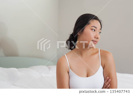 Korean woman wearing camisole sitting on bed corner in lit bedroom with white bedding, copy space Korean woman wearing camisole sitting on bed corner in lit bedroom with white bedding, copy space 135407743