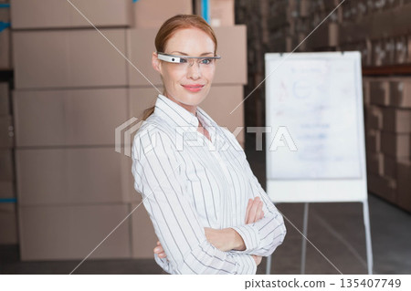 Mid adult woman standing arms crossed in warehouse wearing smart glasses amid boxes and whiteboard Mid adult woman standing arms crossed in warehouse wearing smart glasses amid boxes and whiteboard 135407749