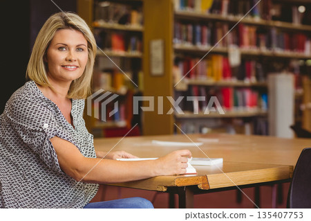 Middle-aged woman holding open book, sitting at library table with laptop and papers, copy space 135407753