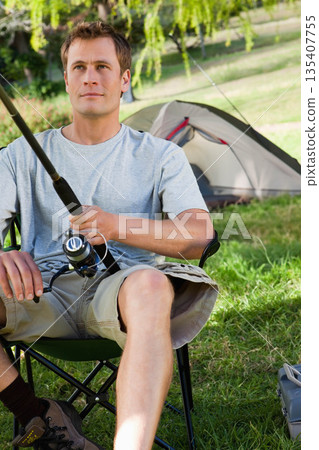 Man holding fishing rod while sitting on foldable camping chair at grassy campsite near dome tent Man holding fishing rod while sitting on foldable camping chair at grassy campsite near dome tent 135407755