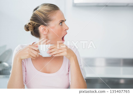 Woman holding white ceramic mug and looking surprised at kitchen counter near dish rack Woman holding white ceramic mug and looking surprised at kitchen counter near dish rack 135407765