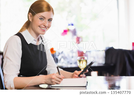Red-haired woman sitting at cafe table writing in notebook while holding smartphone with wine glass Red-haired woman sitting at cafe table writing in notebook while holding smartphone with wine glass 135407770