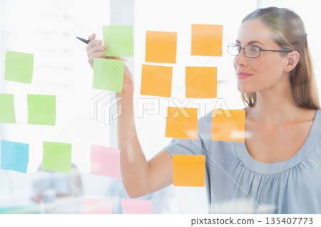 Mid adult female moving sticky note on glass board in meeting room with pen, copy space 135407773