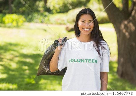 Asian woman volunteering in park clearing wearing volunteer t-shirt and holding black trash bag Asian woman volunteering in park clearing wearing volunteer t-shirt and holding black trash bag 135407778
