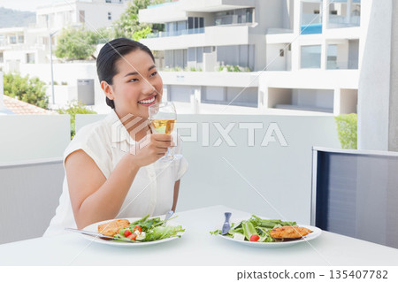Korean woman sitting at balcony table sipping drink from stemmed glass with salad, copy space Korean woman sitting at balcony table sipping drink from stemmed glass with salad, copy space 135407782
