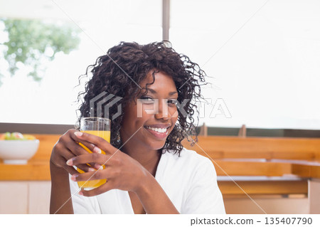 African American woman sitting at wooden table in bright kitchen holding tall glass of orange juice African American woman sitting at wooden table in bright kitchen holding tall glass of orange juice 135407790