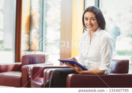 Female business professional holding blue tablet sitting on leather armchair in lobby, copy space 135407795