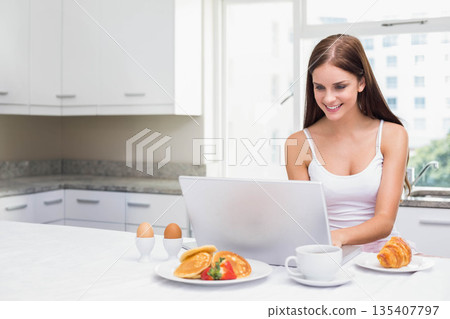 Woman typing on laptop at modern kitchen island with pancakes topped with strawberries eggs mug Woman typing on laptop at modern kitchen island with pancakes topped with strawberries eggs mug 135407797
