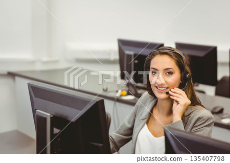 Female call center operator in grey blazer answering calls at office desk with headset, copy space Female call center operator in grey blazer answering calls at office desk with headset, copy space 135407798