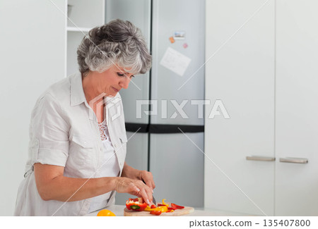 Senior woman slicing yellow and red bell peppers on cutting board in white kitchen, copy space Senior woman slicing yellow and red bell peppers on cutting board in white kitchen, copy space 135407800