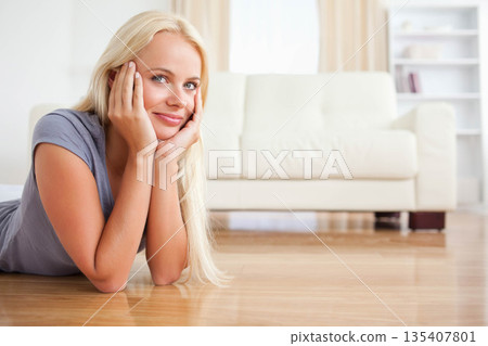 Woman reclining on hardwood floor in living room with white sofa, curtains and shelving, copy space 135407801