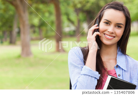 Smiling woman holding notebook under arm while talking on smartphone in park, copy space 135407806