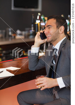 Man wearing gray suit sitting at bar counter holding smartphone and writing on papers with pen 135407816