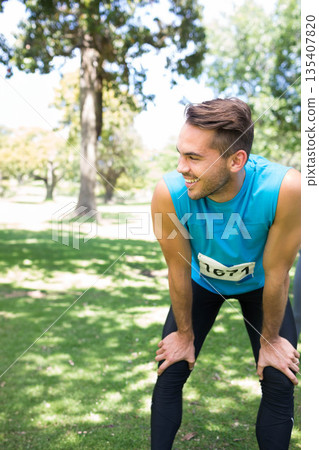 Male runner pausing on grass in park wearing blue shirt and bib 1671, copy space Male runner pausing on grass in park wearing blue shirt and bib 1671, copy space 135407820