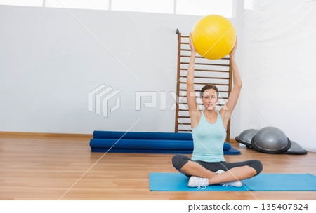 Woman lifting yellow stability ball overhead on blue mat near wooden stall bars in fitness studio 135407824