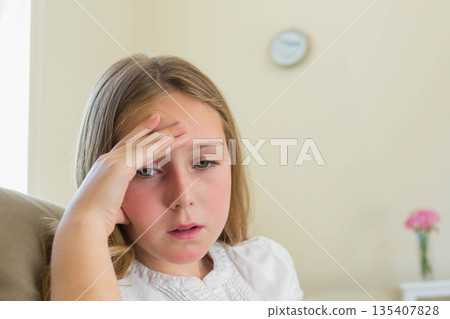 Teenage girl sitting in armchair at home holding forehead beneath clock beside pink flowers Teenage girl sitting in armchair at home holding forehead beneath clock beside pink flowers 135407828