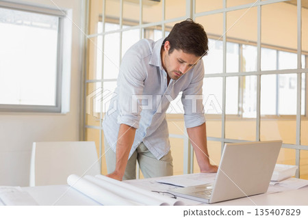 Man in button-down shirt reviewing blueprints on white desk with silver laptop at office 135407829