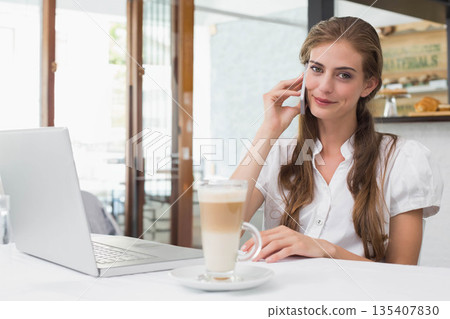 Woman talking on smartphone while typing on laptop at coffee shop with latte, pastries, copy space Woman talking on smartphone while typing on laptop at coffee shop with latte, pastries, copy space 135407830