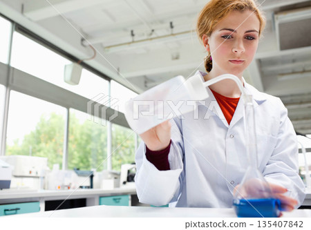 Female scientist holding white wash bottle, pouring into blue beaker at lab bench, copy space 135407842