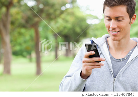 Adult man standing in park holding smartphone and wearing gray hoodie with backpack, copy space 135407845