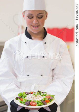 Female chef holding plate of green salad with chicken and purple basil in stainless steel kitchen 135407847