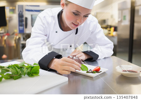Female chef garnishing chocolate dessert wearing white uniform with mint, strawberry in kitchen Female chef garnishing chocolate dessert wearing white uniform with mint, strawberry in kitchen 135407848