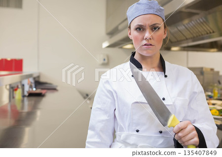 Female chef in chef uniform standing in kitchen holding large knife near red order boxes 135407849