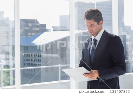Standing male business professional in suit and tie holding tablet in high-rise office, copy space Standing male business professional in suit and tie holding tablet in high-rise office, copy space 135407853