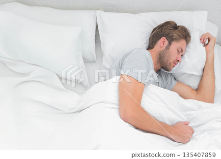 Mid adult male lying in bedroom bed wearing gray shirt, resting on white bedding, copy space 135407859