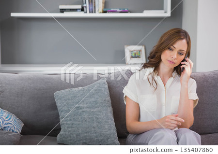 Woman sitting on couch in living room holding smartphone to ear by floating shelf, copy space 135407860