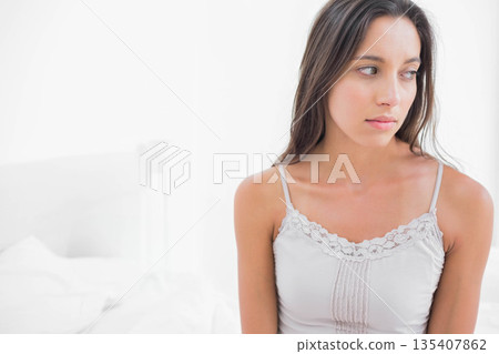 Woman sitting on edge of bed in bright white bedroom wearing lace-trimmed camisole, copy space 135407862