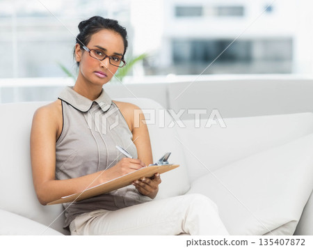 Asian woman writing notes on clipboard while sitting on modern white sofa in bright office lounge 135407872