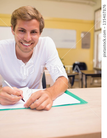 Male student studying at light wood desk in classroom holding pen above paper on green folder Male student studying at light wood desk in classroom holding pen above paper on green folder 135407876