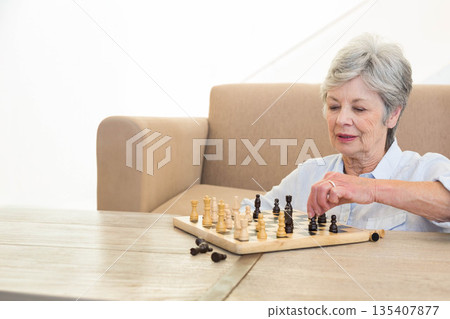 Senior woman focusing on chessboard while playing chess on wooden table in living room, copy space Senior woman focusing on chessboard while playing chess on wooden table in living room, copy space 135407877