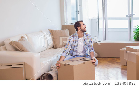 Man looking toward balcony doors while sitting on cardboard moving box in living room Man looking toward balcony doors while sitting on cardboard moving box in living room 135407885