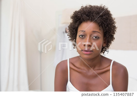 African American woman sitting on bed wearing camisole by beige headboard thinking in bedroom African American woman sitting on bed wearing camisole by beige headboard thinking in bedroom 135407887