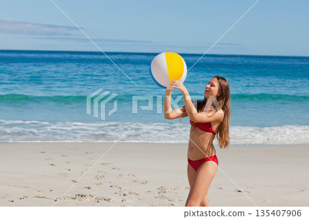 Woman standing wearing red bikini at shoreline beach holding beach ball above head, copy space 135407906