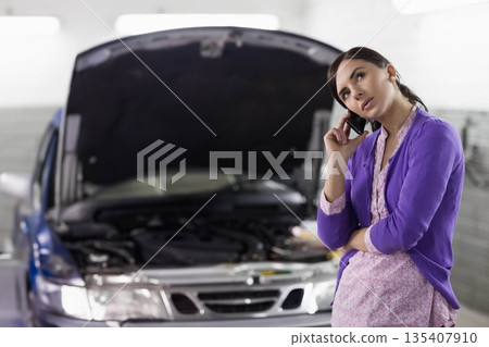 Adult female using smartphone beside silver sedan with engine components in garage, copy space 135407910