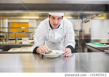 Male chef wearing chef jacket leaning over countertop in kitchen, adding garnishes to salad bowl Male chef wearing chef jacket leaning over countertop in kitchen, adding garnishes to salad bowl 135407913