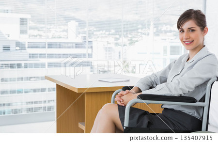 Businesswoman sitting in wheelchair at desk with notebook in office overlooking skyline, copy space Businesswoman sitting in wheelchair at desk with notebook in office overlooking skyline, copy space 135407915