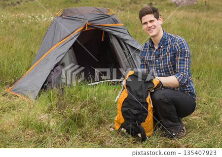 Man kneeling next to gray orange dome tent in grassy meadow organizing backpack gear, copy space 135407923