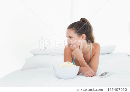 Woman reclining on white bedspread in turquoise tank top, reaching for popcorn bowl near remote Woman reclining on white bedspread in turquoise tank top, reaching for popcorn bowl near remote 135407930